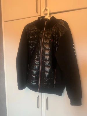 Svart pufferjacka från Tommy Hilfiger - Säljer en svart pufferjacka från Tommy Hilfiger med glansig quiltad framsida och mjuka ärmar. Jackan har dragkedja framtill, huva och logga på bröstet samt stort tryck på ena ärmen. Perfekt för dig som vill ha en stilren och sportig look.