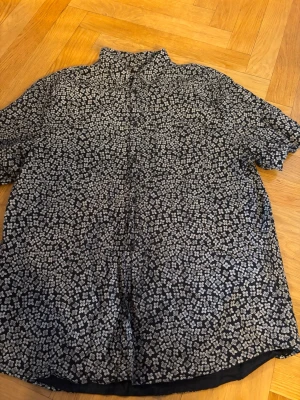 Blommig kortärmad skjorta från H&M - Kortärmad skjorta från H&M i regular fit, svart botten med tätt vitt blommönster. Skjortan har klassisk krage och knäppning framtill. Perfekt för dig som vill sticka ut lite extra med ett coolt mönster.