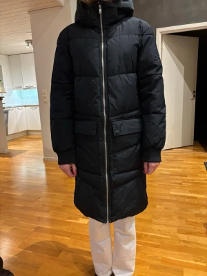Svart lång pufferjacka Everest storlek 170 - Lång svart pufferjacka med huva. Storlek 170 men passar även vuxen xs/s. Vid fickorna finns reflexer som går att vika in och fram igen vid behov. 