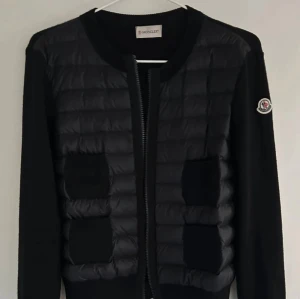 Svart Moncler cardigan  - Säljer en svart pufferjacka från Moncler med quiltad front och stickade ärmar. Jackan har rund hals, två fickor framtill och dragkedja. Moncler-logga på ärmen. Perfekt för dig som vill ha en stilren och sportig look.