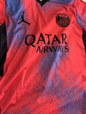 PSG Mbappé matchtröja Nike röd/blå - Säljer en Paris Saint-Germain matchtröja med Mbappé 7 på ryggen. Tröjan är röd med blått stänkmönster, svart Nike-logga och PSG-märke på bröstet. Tillverkad i lätt polyester med svart V-ringad krage och Qatar Airways-tryck på framsidan.