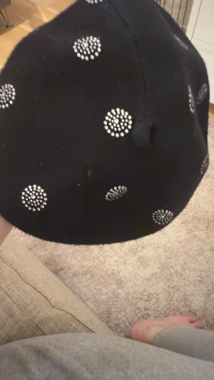 Svart basker med prickiga nitar - Säljer en svart basker dekorerad med silvriga nitar i prickmönster. Klassisk rund form och mjukt material som känns ulligt. Perfekt accessoar för att lyfta din outfit med lite extra edge.