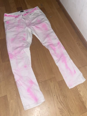 Vita jeans med rosa tie dye, strl 34 - Unika vita jeans med rosa tie dye-effekt. Klassisk femficksmodell med knapp och dragkedja framtill. Byxorna har raka ben och är tillverkade i mjuk bomull. Perfekta för dig som vill sticka ut med färg och kreativ stil.