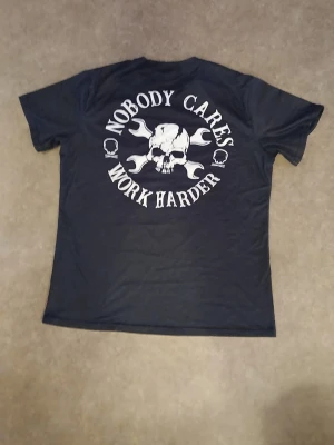 Svart t-shirt med tryck, storlek L - Svart t-shirt från Temu i storlek L med stort vitt tryck på ryggen: döskalle, korslagda skiftnycklar och texten 'Nobody cares, work harder'. Tillverkad i mjukt material med 95% polyester och 5% spandex. Klassisk rund hals och korta ärmar.
