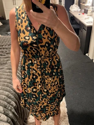 Leopardmönstrad ärmlös klänning med snörning - Säljer en cool leopardmönstrad klänning i beige, svart och grönt. Klänningen är ärmlös, har v-ringad hals och knappar framtill. Midjan kan justeras med snörning för en snygg passform. Perfekt för dig som gillar djurmönster och vill sticka ut.