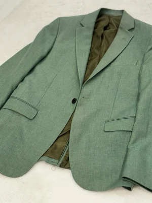 Grön kavaj med klassisk passform - Snygg grön kavaj med klassisk krage och en knapp framtill. Fodrad insida i olivgrönt tyg och två fickor med lock. Perfekt för dig som vill ha en stilren look med lite färg. Passar till både jeans och kostymbyxor.