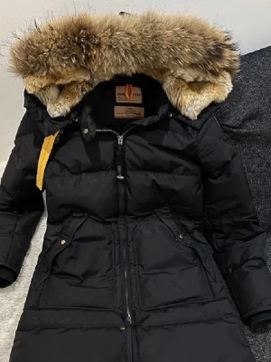 Svart Parajumpers dunjacka med päls - Säljer en svart lång dunjacka från Parajumpers med stor fluffig päls runt huvan och snygga detaljer som gul rem och patch på ärmen. Jackan har dragkedja framtill, stora fickor och är perfekt för kalla vinterdagar. Klassisk och tidlös modell som håller dig varm.