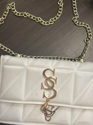 Vit quiltad axelväska från Guess - Säljer en snygg vit quiltad axelväska från Guess med guldfärgad kedja och logga framtill. Väskan har elegant design med triangulärt mönster och dekorativt hängsmycke. Perfekt för dig som vill ha en trendig accessoar med lyxig känsla.