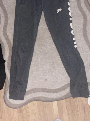 Mörkgrå Nike mjukisbyxor XS - Mörkgrå mjukisbyxor från Nike i storlek XS. Byxorna har resår i bensluten, vit Nike-logga på höften och stor vit text 'JUST DO IT.' längs ena benet. Tillverkade i mjukt material som passar perfekt för chill eller träning.