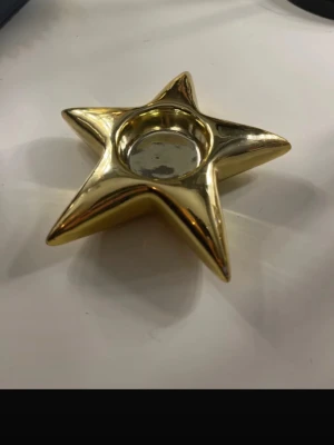 Stjärnformad ljusstake i guldfärgad - Dekorativ ljusstake formad som en stjärna med en elegant guldfärgad yta. Passar perfekt som stämningshöjande inredningsdetalj på bord, hylla eller fönsterbräda. Ljusstaken har en modern och stilren design som gör sig lika bra till vardags som vid högtider. Observera att lite av färgen har försvunnit på vissa partier, vilket syns vid närmare granskning och ger ett lätt patinerat intryck.