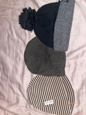 Tre mössor i olika färger och mönster - Säljer tre mössor: en svart och grå stickad mössa från Everest med tofs, en melerad grågrön stickad mössa och en beige och svart randig mössa från Lilla-Luv. Perfekta för kalla dagar och snygga till alla outfits.Är barnmössor!