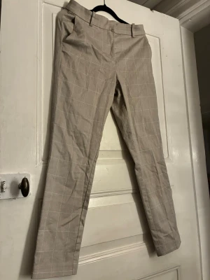 Beige rutiga kostymbyxor från H&M, storlek 38 - Säljer ett par beige kostymbyxor från H&M med vitt rutmönster. Byxorna har bälteshällor, raka ben och två fuskfickor bak. Materialet känns som bomull eller polyester och de har en klassisk, stilren look. Perfekta till skjorta eller stickad tröja.
