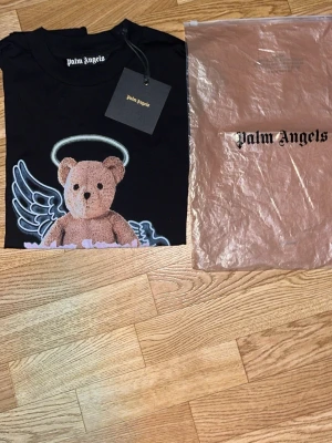 Palm Angels svart t-shirt med nalle - Svart t-shirt från Palm Angels med tryck av en nalle med gloria och vingar på bröstet. Klassisk rund hals och korta ärmar. T-shirten är i bomull och har en cool streetwear vibe. Perfekt för dig som gillar statement-plagg och snygga prints.