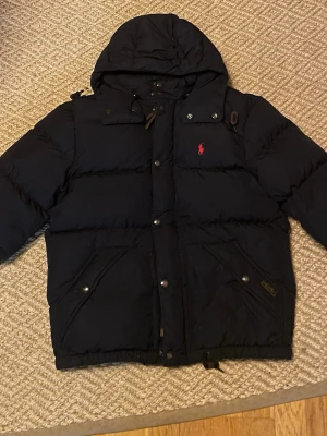 Marinblå pufferjacka från Polo Ralph Lauren - Säljer en marinblå pufferjacka från Polo Ralph Lauren i storlek S som är i riktigt bra skick. Nypris på jackan är runt 5 tusen. Några av metalknapparna har tappat lite färg men det är det ända felet Inga hål eller liknande. Skriv för mer info. Jackan har huva, dragkedja och knappar framtill samt två stora fickor. Klassisk röd logga på bröstet och snygga detaljer. Perfekt för kalla dagar!