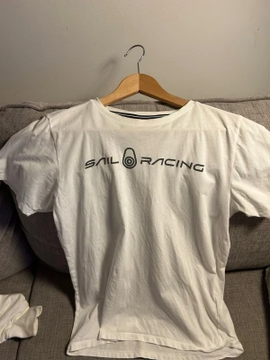 Vit Sail Racing t-shirt i bomull - Vit t-shirt från Sail Racing med svart logga tryckt över bröstet. Klassisk rund halsringning med svart-vit randig detalj på insidan av kragen. Skön och enkel modell i mjuk bomull, perfekt till jeans eller shorts. Nyskick. Använd 1 gång 
