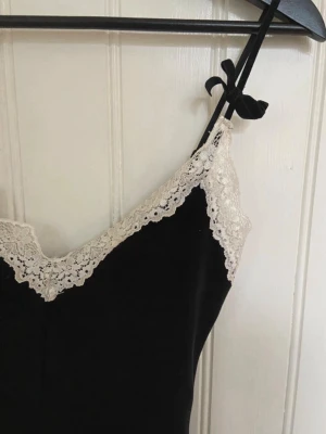 Svart sammetsklänning med spets från Motel Rocks - Supersöt svart sammetsklänning från Motel Rocks med smala axelband och dekorativa rosetter. V-ringad med vit spets längs urringningen som ger en romantisk vibe. Endast använd en gång!💕