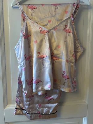 Pyjamas med flamingomönster - Fin pyjamas i glansigt material med champingerosa bas och rosa flamingos samt små rosa hjärtan. Toppen har smala axelband och byxorna är breda för en avslappnad passform, perfekt för en lyxig chill look. 