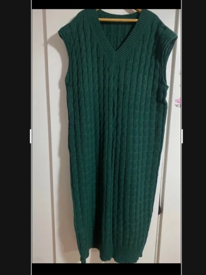 Grön stickad långklänning med v-ringning - Mysig grön stickad långklänning med v-ringad hals och ärmlös design. Klänningen har ett snyggt kabelstickat mönster och ribbade kanter upptill och nertill. Perfekt för lager-på-lager och chill dagar.