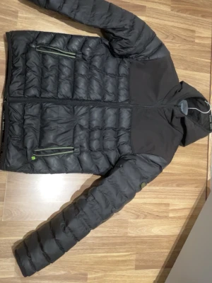 Svart pufferjacka från Hugo Boss - Säljer en svart pufferjacka från Hugo Boss med huva och gröna detaljer på dragkedjorna. Jackan har quiltad design, lång ärm och mjukt foder på insidan. Stort Hugo Boss-tryck på ryggen och logga på bröstet. Perfekt för kalla dagar och riktigt snygg streetstil.