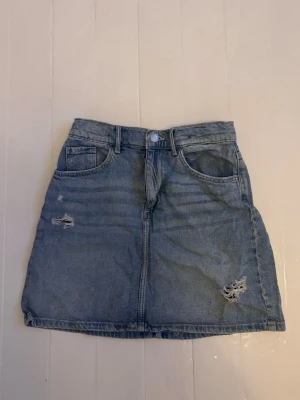 Blå jeanskjol med slitningar - Säljer en blå jeanskjol i klassisk modell med råa slitningar framtill. Kjolen har fem fickor, bälteshällor och knappstängning. Perfekt för dig som gillar en avslappnad och trendig stil.