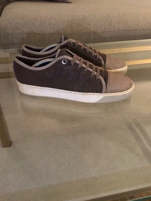 Lanvins, bruna sneakers  - Säljer snygga bruna sneakers från Lanvin med beige mockadetaljer på tå och runt kanten. Skorna har grå sula som ger en clean look. Ordinarie sula ej kvar men annars i bra skick. Perfekta för dig som vill ha en elegant och klassisk stil. Obs ändats skorna. 
