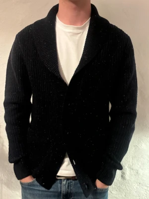 Svart ribbad kofta med sjalkrage - Mysig svart ribbad kofta med bred sjalkrage. Koftan är långärmad och har en klassisk, lite loose passform. Perfekt för lager på lager och funkar till både jeans och t-shirt. Materialet känns tjockt och stickat, vilket gör den extra skön under kyliga dagar. Märke, Selected Home, nytt pris 499 vårt pris 199kr. Hör av dig vid funderingar 