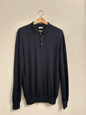 Mörkblå pikétröja i merinoull från John Henric - Klassisk mörkblå långärmad pikétröja från John Henric i mjuk merinoull. Tröjan har krage, tre knappar framtill och ribbade muddar vid ärmslut och nederkant. Perfekt för dig som gillar stilrena och tidlösa plagg.