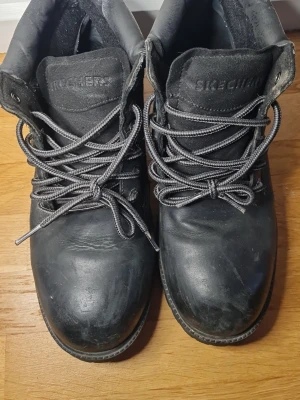 Svarta boots från Skechers i skinn - Tuffa svarta boots från Skechers i skinn med grov sula och kraftiga snören. Klassisk rund tå och metallhål för snörningen. Perfekta för dig som vill ha en robust och stilren look till höst och vinter.