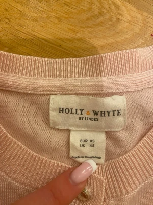 Ljusrosa kofta från Holly & Whyte XS - Supersöt ljusrosa kofta från Holly & Whyte by Lindex i storlek XS. Klassisk rund halsringning, långa ärmar och knappar framtill. Tunn och mjuk kvalitet, perfekt för lager på lager. Liten metallbricka med logga vid nederkanten.