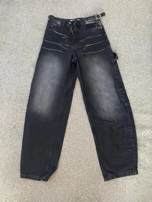 Barrel svarta jeans - Säljer ett par svarta baggy barrel jeans från Bershka som är oanvända. Byxorna har bred passform, fickor och bältesdetalj i midjan. Perfekta för dig som gillar streetwear och vill sticka ut lite extra.