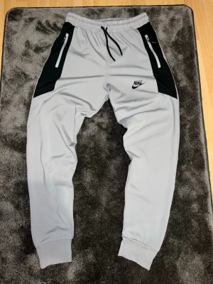 Nike grå träningsbyxor med svarta detaljer - Nike träningsbyxor i ljusgrått med svarta paneler vid fickorna och svart Nike-logga på låret. Byxorna har dragkedjefickor, resår och snörning i midjan samt muddar vid bensluten. Materialet är syntet och passar perfekt för träning. Står att det är L men känns som S passar också M! 