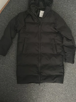 Svart lång pufferjacka Jack & Jones - Säljer en svart lång pufferjacka från Jack & Jones i storlek S. Jackan har huva, dragkedja framtill och stora fickor med tryckknapp. Perfekt för kalla dagar och riktigt skön att bära. Enkel och stilren design som funkar till det mesta.