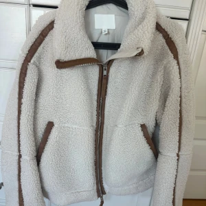 Beige teddyjacka från H&M  - Säljer en beige teddyjacka från H&M i storlek XS. Jackan har bruna detaljer längs dragkedjan, fickorna och ärmarna, samt en hög krage. Materialet är mjuk fleece med syntet och jackan har två fickor framtill och en rejäl dragkedja.