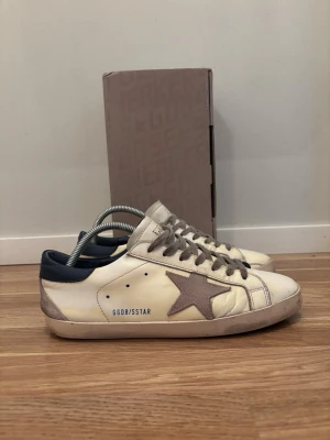 Golden Goose vita sneakers med stjärna - Säljer ett par klassiska Golden Goose sneakers i vitt skinn med grå mockadetaljer och stjärna på sidan. Skorna har grå snörning, mörkblå häl och beige sula. Perfekta för dig som gillar en avslappnad och trendig look. Lite tecken på slititage arnas väldigt schyssta. 