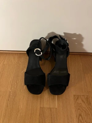 Svarta sandaler med klack från Attitude - Säljer ett par svarta sandaler från Attitude med bred rem över tårna och justerbart spänne i silver. Skorna har en chunky hög klack och är tillverkade i mockaimitation. Perfekta för dig som vill ha en stilren och enkel look.