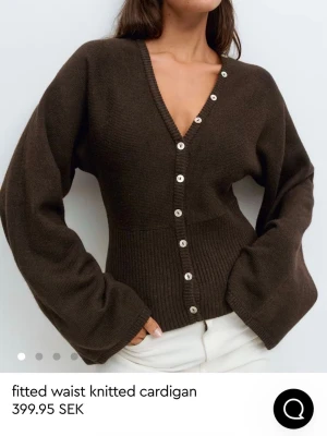 Brun stickad kofta med knappar - Mörkbrun stickad kofta med v-ringning och markerad midja. Koftan har långa, vida ärmar och stängs med en rad vita knappar framtill. Perfekt för lager på lager under kyligare dagar. Endast använd en gång