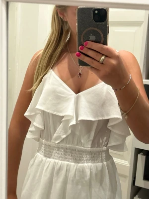 Vit omlottklänning med volang - Säljer en vit kortklänning med omlott-design och bred volang upptill. Klänningen har smala axelband och en smockad midja som ger en snygg passform. Materialet är lätt och luftigt, perfekt för varma dagar. Stilren och fräsch look.