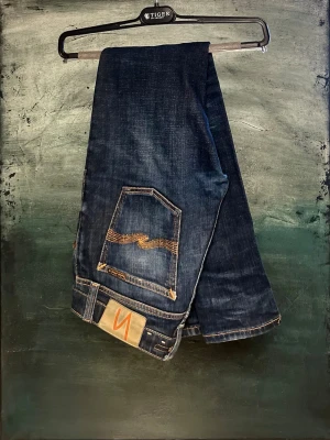 Nudie Jeans Herr - Säljer ett par mörkblå slim fit jeans från Nudie Jeans med klassiska orange sömmar och vågig brodyr på bakfickorna. Jeansen har normal midja och är tillverkade i slitstarkt bomullsdenim. Perfekta för dig som gillar en lite smalare passform och stilren look.