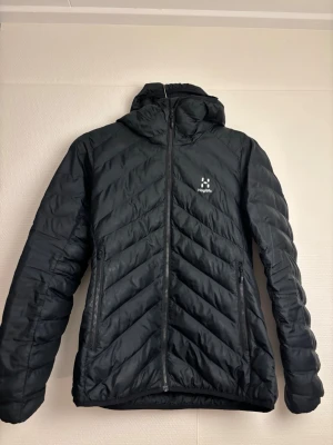 Svart pufferjacka från Haglöfs - Säljer en svart pufferjacka från Haglöfs med huva och dragkedja framtill. Jackan har snygga diagonala sömmar på framsidan och raka sömmar på ryggen. Tillverkad i syntetmaterial och har två praktiska fickor med dragkedja. Perfekt för kalla dagar.