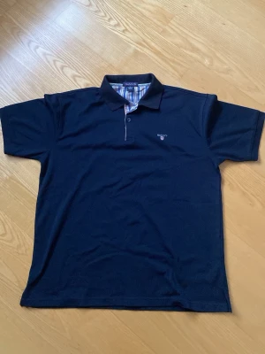 Marinblå pikétröja från GANT XL - Snygg marinblå pikétröja från GANT i storlek XL. Klassisk krage, korta ärmar och broderad logga på bröstet. Insidan av kragen har ett färgglatt randigt tyg i blått, rött och vitt. Perfekt till en clean och stilren look.