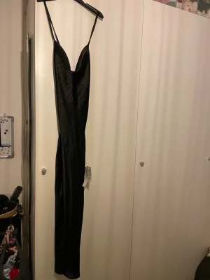 Svart långklänning i satin - Säljer en svart långklänning med smala axelband och snygg drapering framtill. Klänningen har snörning i ryggen för en justerbar passform och är gjord i ett glansigt satin material som ger en lyxig känsla. Perfekt för speciella tillfällen. Aldrig använd bara testad med prislappen kvar. Pris kan diskuteras.