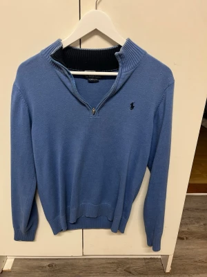 Blå stickad tröja från Ralph Lauren - Säljer en blå stickad tröja från Ralph Lauren med halv dragkedja och ribbad krage. Tröjan har lång ärm och den klassiska broderade loggan på bröstet. Perfekt för lager-på-lager och chillare dagar.