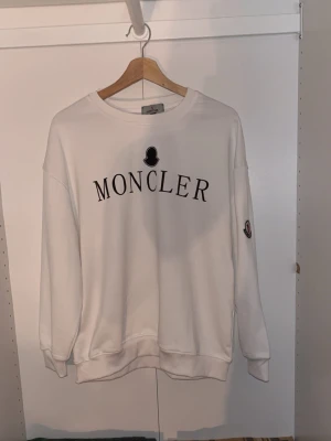Vit sweatshirt från Moncler - Säljer en vit sweatshirt från Moncler med svart logotyptryck på bröstet och ett broderat Moncler-märke på ärmen. Tröjan har rund hals, ribbade muddar och är långärmad. Perfekt för dig som gillar stilrena och exklusiva plagg.