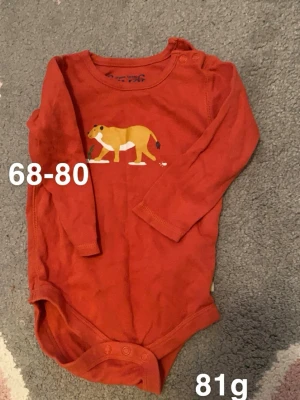 Orange långärmad body med lejon - Superfin orange långärmad body med tryck av ett gult lejon framtill. Bodyn har praktiska knappar på axeln och i grenen för enkel påklädning. Perfekt för små äventyrare som gillar djurmotiv.🌸 Två små fläckar. storlek 68-80, upplever som storlek 74