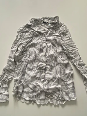 Randig vit skjorta  - Säljer en vit skjorta från H&M med svarta vertikala ränder. Skjortan har klassisk krage, knäppning framtill och långa ärmar. 