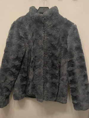 Grå jacka med dragkedja, storlek 44/46  - Mysig grå pälsjacka med hög krage och dragkedja framtill. Jackan har ett fluffigt och mjukt material med struktur i ”pälsen” som ger en cool look. Perfekt för dig som vill ha en varm och trendig jacka till höst och vinter. Storlek 44/46.