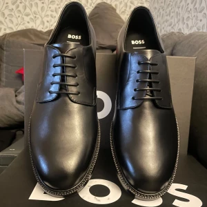 Nya Hugo Boss, strl 42. - Hugo Boss Finskor i storlek 42, tillverkade i svart läder. Skorna har en klassisk design med snörning och är tillverkade i Italien. Sulan har ett mönster för bättre grepp och komfort. Passar för formella och professionella sammanhang. Helt nya i originalkartong. Pris kan diskuteras vid snabb affär.