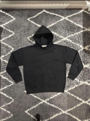 Fear of God Essentials hoodie Svart - Svart hoodie med huva och känguruficka. Stilren design med diskret Essentials-logga på bröstet. Mjuk och bekväm passform, perfekt för vardagsbruk. Jag väger 70KG och är 180CM och den passar mig perfekt.