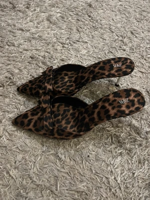 Leopardmönstrade pumps från Zara - Säljer ett par snygga pumps från Zara med leopardmönster i brunt och svart. Skorna har spetsig tå, låg klack och en dekorativ rosett framtill. Perfekta för dig som vill sticka ut med ett djurmönstrat statement. Materialet känns mjukt och lyxigt.