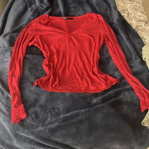 Röd spetsdetaljerad topp från Bik Bok - Säljer en röd långärmad topp från Bik Bok i storlek L. Toppen har v-ringning med spetsdetaljer och spets vid ärmsluten. Materialet är mjukt och stretchigt, vilket ger en bekväm passform. Perfekt för dig som vill ha något snyggt och enkelt.